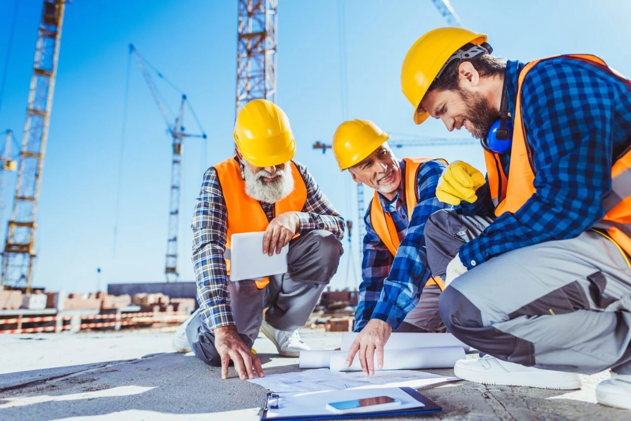 Three-construction-workers-sitting-on-concrete-at-construction-site-discussing-building-plans-e1700584017214